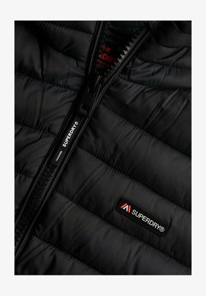 Chaqueta de plumas negra con textura acolchada, que presenta una cremallera negra y un pequeño parche con el logo "Superdry" en rojo y blanco en el pecho.