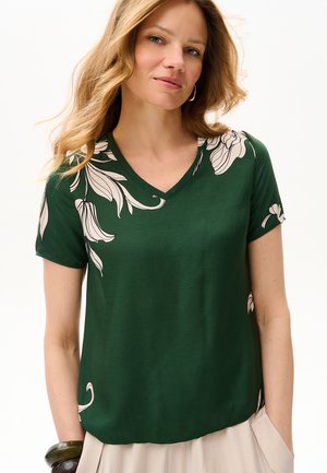 Kobieta w ciemnozielonym topie z krótkim rękawem i dekoltem w serek z beżowymi kwiatowymi wzorami, beżowych spodniach oraz ciemnych bransoletkach na lewym nadgarstku.