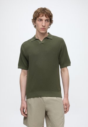 Junger Mann trägt ein olivgrünes, strukturiertes kurzärmeliges Poloshirt und beige Shorts, steht vor einem schlichten weißen Hintergrund.