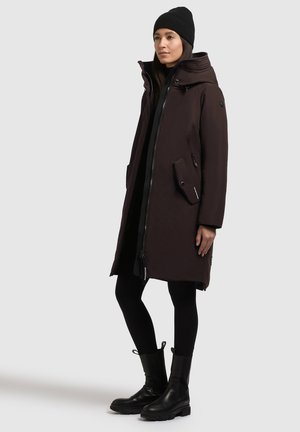 Donkerbruine parka jas met een rits aan de voorkant, een oversized capuchon, twee zijzakken en een gestructureerde afwerking. Gecombineerd met zwarte leggings en enkellaarsjes.