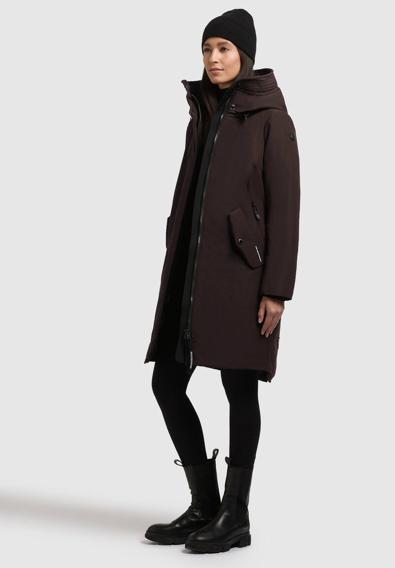 Parka de color marrón oscuro con cremallera frontal, capucha oversized, dos bolsillos laterales y un acabado texturizado. Combinada con leggins negros y botas de tobillo.