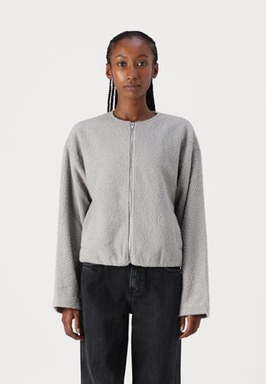 Grau Fleece-Jacke mit Rundhalsausschnitt, Reißverschluss und langen Ärmeln. Weicher Stoff und cropped Passform, kombiniert mit dunklen Jeans.