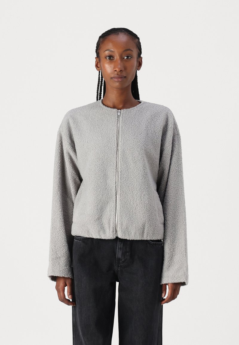 Grå fleecejakke med rund hals, glidelås og lange ermer. Myk tekstur og kort passform, kombinert med mørke jeans.