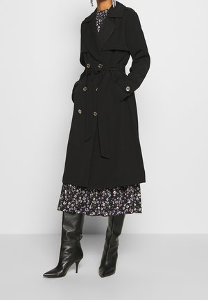 Personne portant un trench-coat noir ceinturé par-dessus une robe fleurie, associé à des bottes noires montantes à talons, debout devant un fond uni.