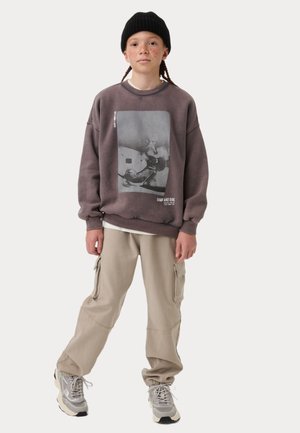 Felpa viola con stampa grafica, pantaloni cargo beige e sneakers grigi. L'outfit presenta una vestibilità rilassata e dettagli di stile casual.