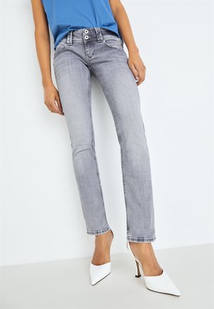 Persoon die lichtgrijze slim-fit jeans, een blauw topje en witte puntige hoge hakken draagt, staand tegen een witte achtergrond.