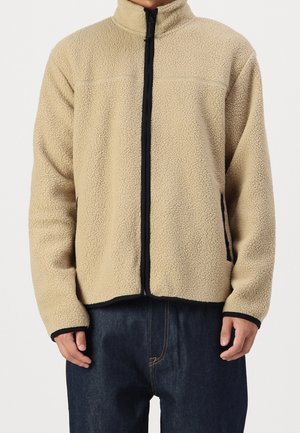 Beige fleecejack met ritssluiting, afgewerkt met zwarte bies, gedragen door een persoon in donkerblauwe spijkerbroek, frontaal beeld van torso en handen.