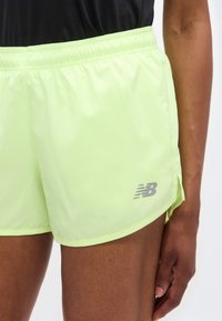 Λαχανί βρακάκι τρεξίματος με γυαλιστερή υφή, ελαστική ζώνη μέσης και πλαϊνές σχισμές. Διαθέτει μικρό γκρι λογότυπο New Balance στο κάτω μέρος.