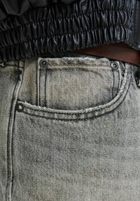 Jean en denim gris clair avec une texture déchirée, dotés d'une poche avant avec quincaillerie en métal et d'une taille élastique noire au-dessus.