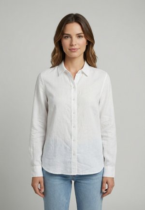 Junge Frau mit schulterlangem braunem Haar, die ein weißes Hemd mit Knöpfen und hellblaue Jeans trägt, steht vor einem einfarbigen grauen Hintergrund.