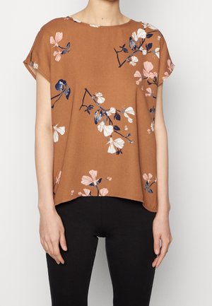 Bruine blouse met korte mouwen en roze en marineblauwe bloemprint, gedragen met een zwarte broek door een staande persoon.
