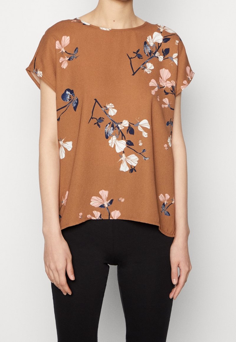 Blouse marron à manches courtes avec imprimés floraux roses et bleu marine portée avec un pantalon noir sur une personne debout.