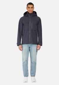 Laineen vedenpitävä takki hupulla, edessä vetoketju, kaksi suurta rintataskua ja vetoketjullinen tasku vasemmalla puolella. Päällä vaaleansiniset farkut.