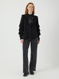 Sort bluse med rysjekanter på ermer og krage, med en sløyfe i sateng. Kombinert med grå vide bukser og svarte sko.