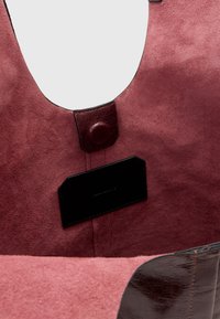 Borsa a tracolla in suede bordeaux con apertura rotonda, chiusura a scatto in pelle e etichetta in pelle nera all'interno. Texture morbida, finitura liscia.