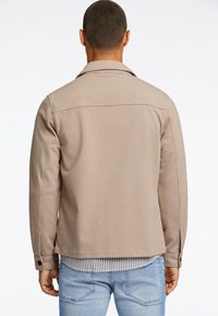 Beige Jacke mit langen Ärmeln, geknöpften Manschetten und einem Kragen. Strukturierter Stoff mit einem dezenten Muster, über einem gestreiften Hemd getragen.