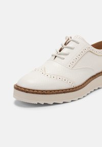 Chaussure oxford en cuir blanc avec une surface texturée, bout rond et perforations décoratives. Dotée de lacets gris et d'une semelle empilée et texturée.