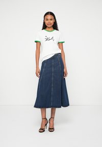 Bijela majica s zelenim obrubom koja sadrži tiskani dizajn, u kombinaciji s dugom traper suknjom strukturiranog A-silhuetnog kroja. Crne sandale s visokom petom.