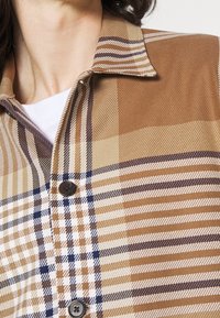 Camisa de botones con un patrón de cuadros marrón y beige con acentos en azul marino. Fabricada en un tejido suave, tiene un cuello con solapas y dos botones.