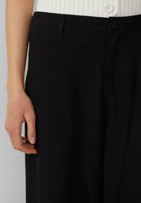 Black fabric pants with a smooth texture, featuring a button closure and a contrasting waistband. Visible stitching detail and side pocket.