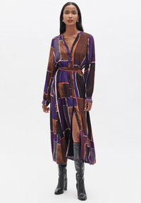 Abito a maniche lunghe con bottoni di colore viola, caratterizzato da un motivo geometrico marrone e arancione. L'abito è dotato di una cintura e di una spacco alto fino alla coscia.