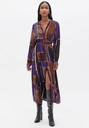 Langarm, doorknopen jurk in paars, met een geometrisch patroon in bruin en oranje. De jurk heeft een riem en een hoge split tot boven de bovenbeen.