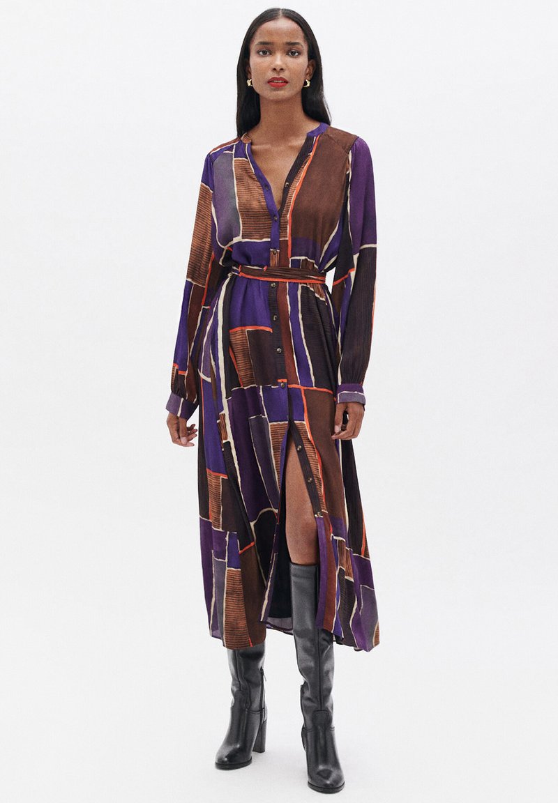 Abito a maniche lunghe con bottoni di colore viola, caratterizzato da un motivo geometrico marrone e arancione. L'abito è dotato di una cintura e di una spacco alto fino alla coscia.
