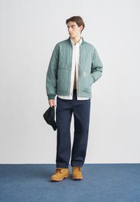 Miętowa pikowana kurtka bomberka z wysokim kołnierzem, przednimi kieszeniami; noszona na białej koszuli, granatowych spodniach i brązowych zamszowych butach. W ręku czapka czarna.