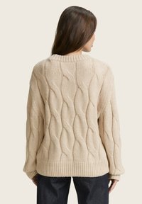 Pull en maille beige avec un motif texturé, à col rond et avec des poignets et un ourlet côtelés. Porté sur un jean noir.