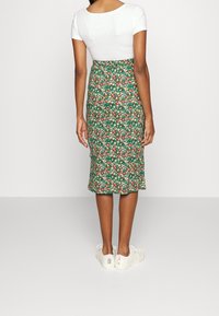 Woman wearing a green floral knee-length skirt, white short-sleeve top, and white sneakers, standing facing a plain wall.