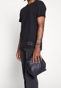 Man med svart t-shirt och mörka jeans som håller en hopvikt svart jacka i handen och visar upp ett handledstatuering, mot en enkel vit bakgrund.