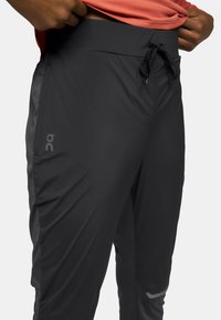 Pantalon de sport noir avec une taille à cordon, présentant des accents sur les panneaux latéraux, un logo réfléchissant et une texture lisse et légère.