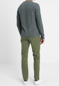 Grön långärmad skjorta med vertikala ränder, ihop med olivgröna byxor. Klädseln har en avslappnad silhuett och vita sneakers.