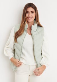 Hellgrüne Steppweste mit hohem Kragen, Rautensteppmuster, durchgehendem Reißverschluss und kombiniert mit einem cremefarbenen Sweater. Glatte Textur.