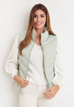 Hellgrüne Steppweste mit hohem Kragen, Rautensteppmuster, durchgehendem Reißverschluss und kombiniert mit einem cremefarbenen Sweater. Glatte Textur.