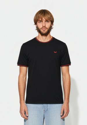 Junger Mann mit hellbraunen Haaren und Bart, der ein schwarzes T-Shirt mit rotem Besatz an Ärmeln und Kragen trägt, kombiniert mit hellblauen Jeans.