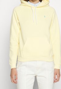 Gul hoodie i mjukt material med en känguruficka, vita snören och en liten logotyp broderad på bröstet. Styler med vita byxor.