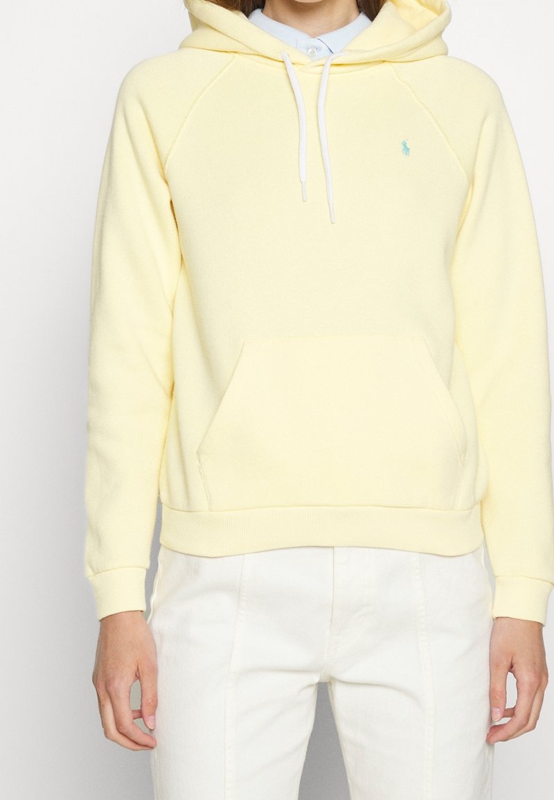 Gul hoodie i mjukt material med en känguruficka, vita snören och en liten logotyp broderad på bröstet. Styler med vita byxor.