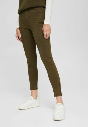 Personne portant un pantalon slim vert olive avec des fermetures éclair aux chevilles et des baskets blanches, debout devant un fond clair et uni.