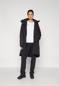 Giacca nera impermeabile con cappuccio, di lunghezza lunga, con zip frontale e grandi tasche; abbinata a una camicia bianca e pantaloni neri.