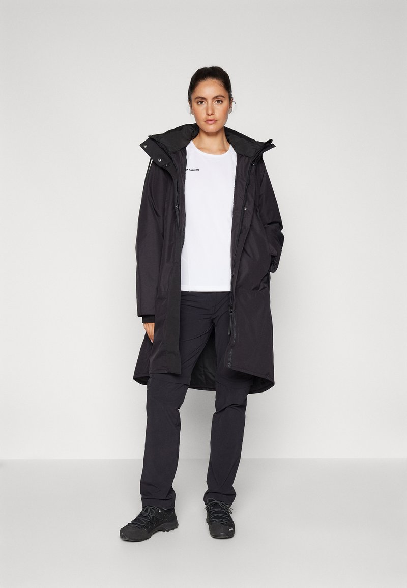 Schwarze wasserdichte Jacke mit Kapuze, langer Schnitt, Frontreißverschluss und großen Taschen; kombiniert mit einem weißen Shirt und schwarzen Hosen.