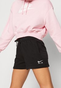Světle růžová zkrácená mikina s bílým logem Nike, v kombinaci s černými šortkami se bílým logem Nike Air, obojí vyrobeno z měkké látky.