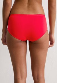 Frau in einem roten, hochgeschnittenen Bikini-Höschen, von hinten vor einem schlichten weißen Hintergrund abgebildet.