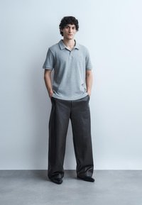 Jeune homme aux cheveux bouclés se tenant contre un mur blanc, portant un polo gris, un pantalon large noir texturé et des chaussures noires, les mains dans les poches.