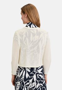 Afgeknotte, crèmekleurige gebreide cardigan met een gestructureerd, open weefontwerp. Beschikt over lange mouwen en een rond decolleté.