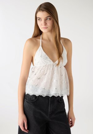 Giovane donna con lunghi capelli castani, che indossa un top bianco ricamato con collo all'americana e pantaloni neri a vita alta, che guarda di lato.