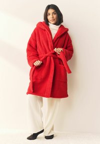 Roter Fleece-Bademantel mit Kapuze, Bindegürtel und zwei Vordertaschen. Weicher, strukturierter Stoff; kombiniert mit cremefarbenen Hosen und dunklen Socken.