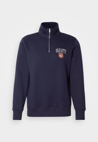 Bluza de navy cu guler înalt, fermoar pe față și manșete cu nervuri. Include logo-ul "GANT" și un emblema cu scut pe piept.