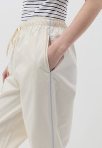 Pantalones color crema con un corte relajado, que cuentan con una cinturilla elástica y bolsillos laterales. Acento azul a lo largo de las costuras laterales. Textura suave.