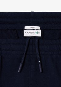 Pantalon de survêtement bleu marine avec une taille à cordon. Comprend une étiquette portant les mentions "LACOSTE" et "REGULAR FIT". Le matériau semble doux et texturé.